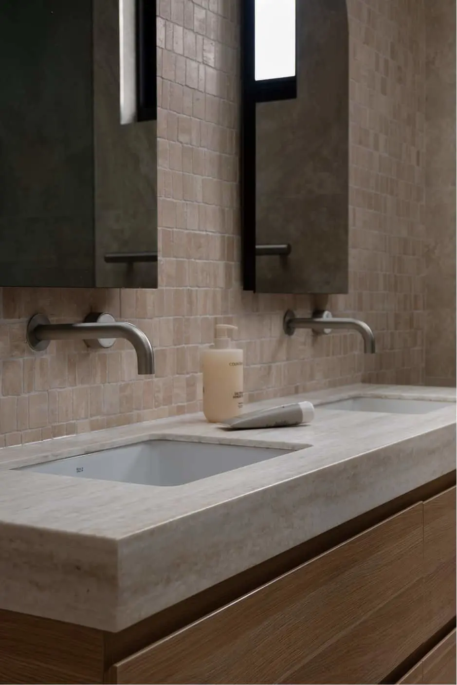 Modern bathroom vanity with stone countertop, wall-mounted faucets, and beige tile backsplash with mirror above.