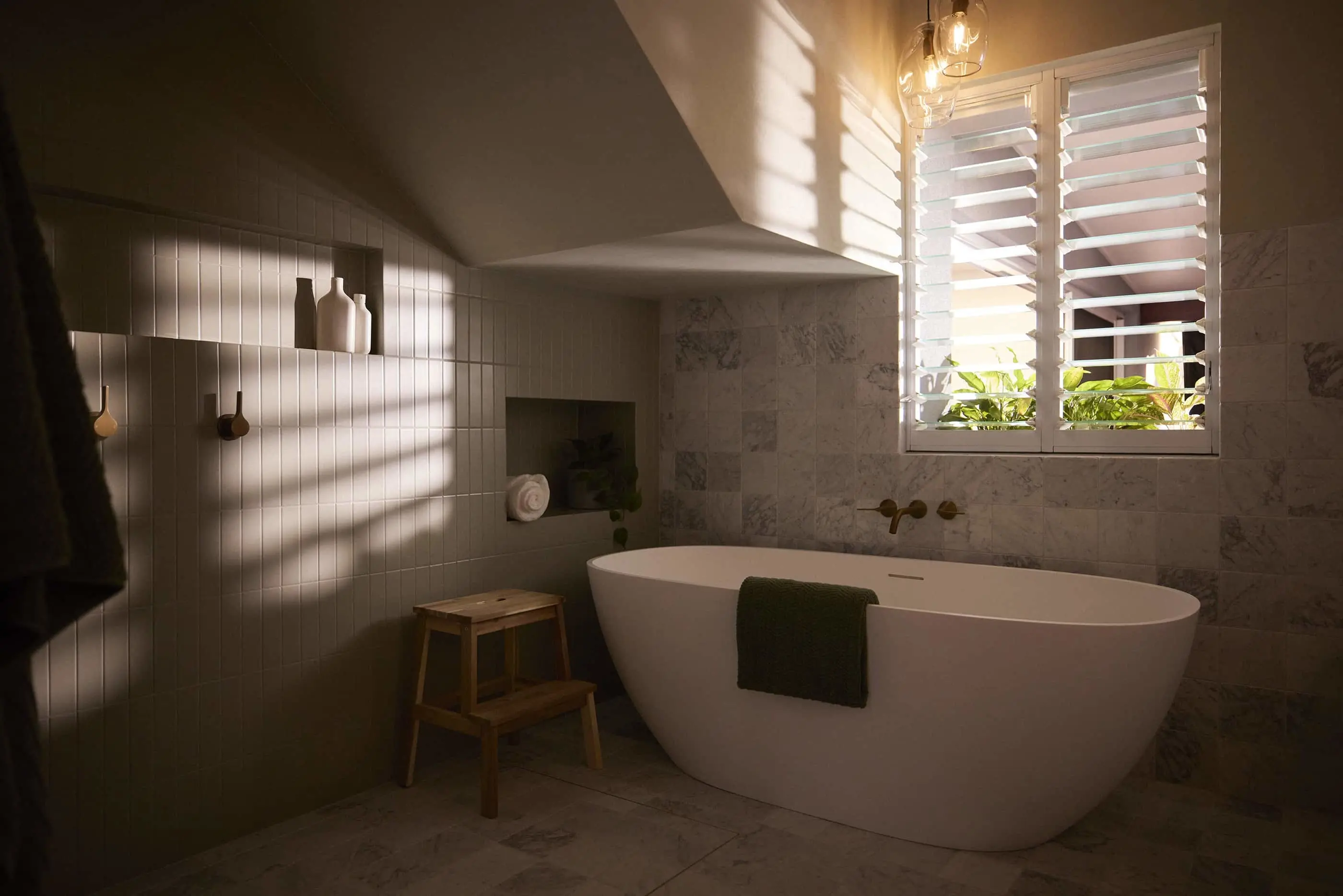 Modern bathroom with white freestanding tub, marble tiles, wooden stool, and sunlight streaming through shuttered window.