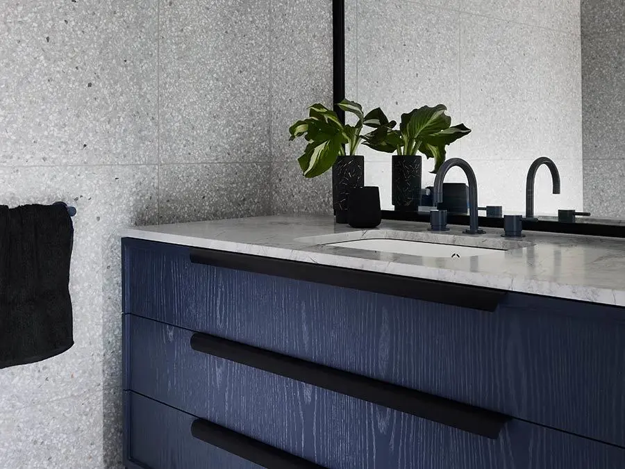 Modern bathroom vanity with navy blue cabinets, marble countertop, black fixtures, and small green plants against gray walls.