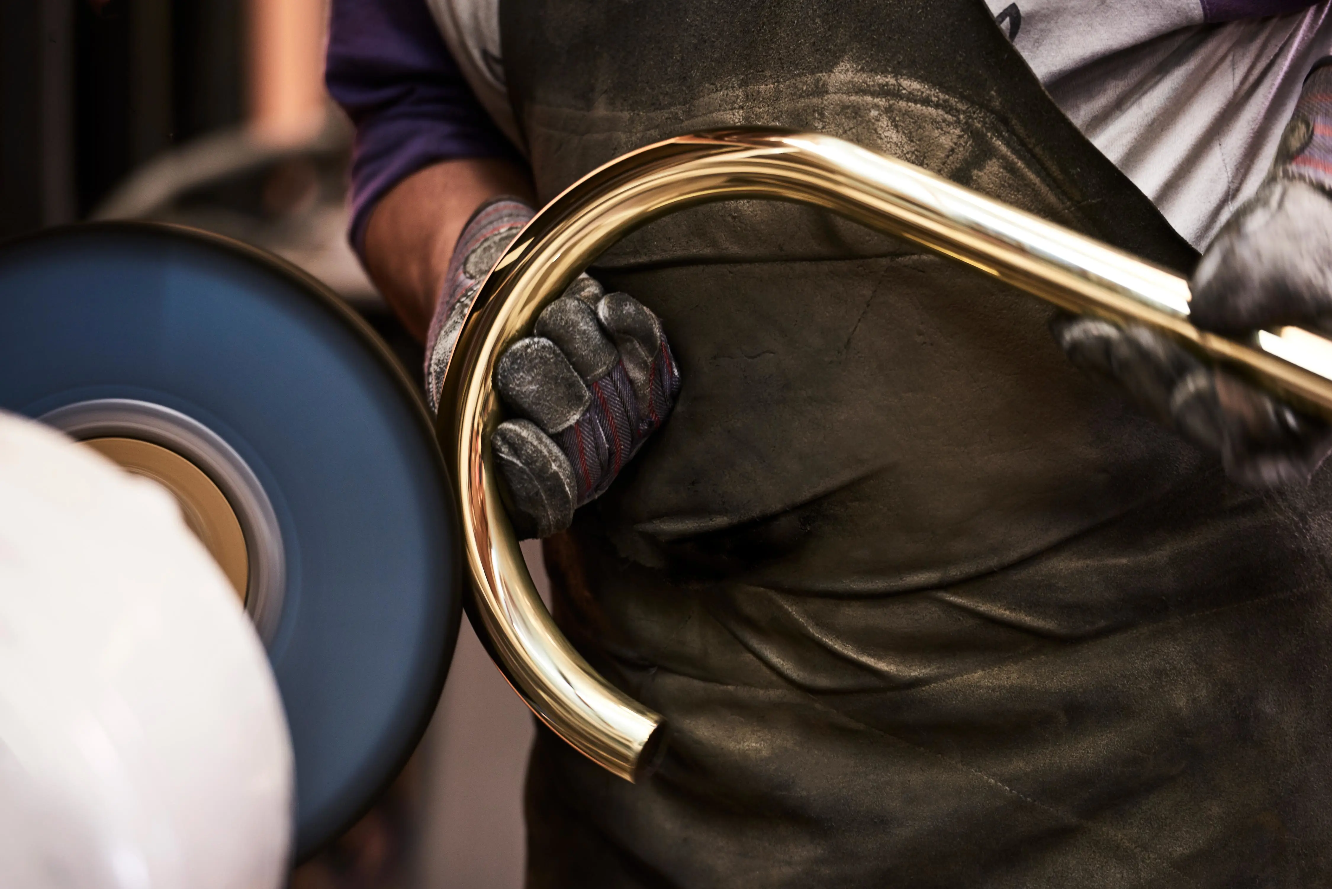 A gloved worker in a dark apron polishes a brass instrument tube against a rotating buffing wheel.