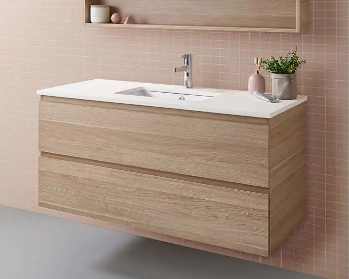 Modern bathroom vanity with light wood cabinet, white countertop, and pink tiled wall featuring small plant decoration.