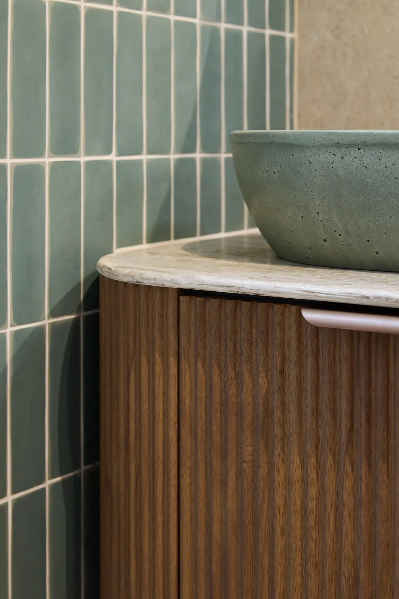 Green concrete bowl on marble countertop with wooden cabinet below, against sage green vertical tile backsplash.