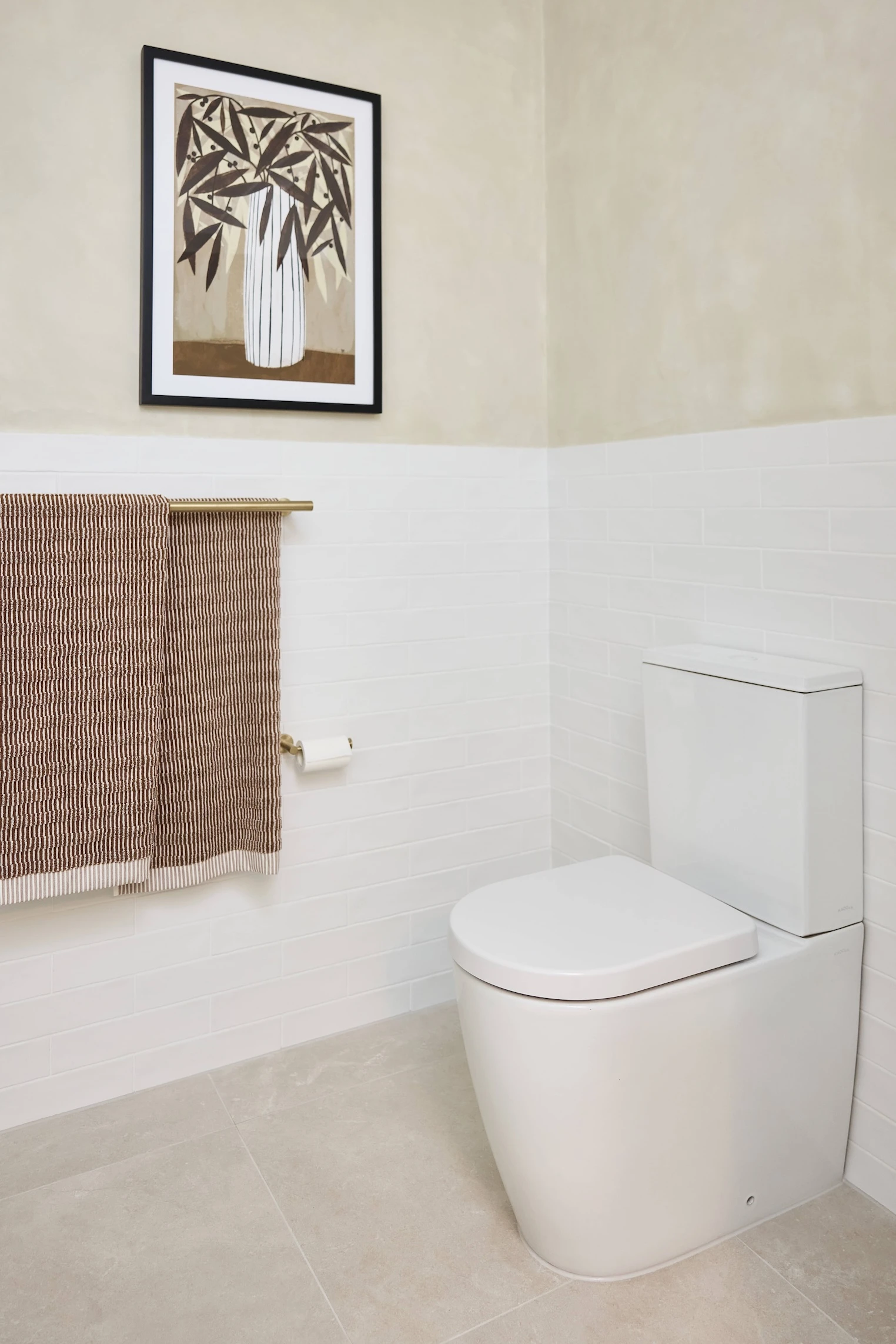 Modern bathroom with white toilet, striped brown towel on brass rail, and framed botanical artwork on beige wall.
