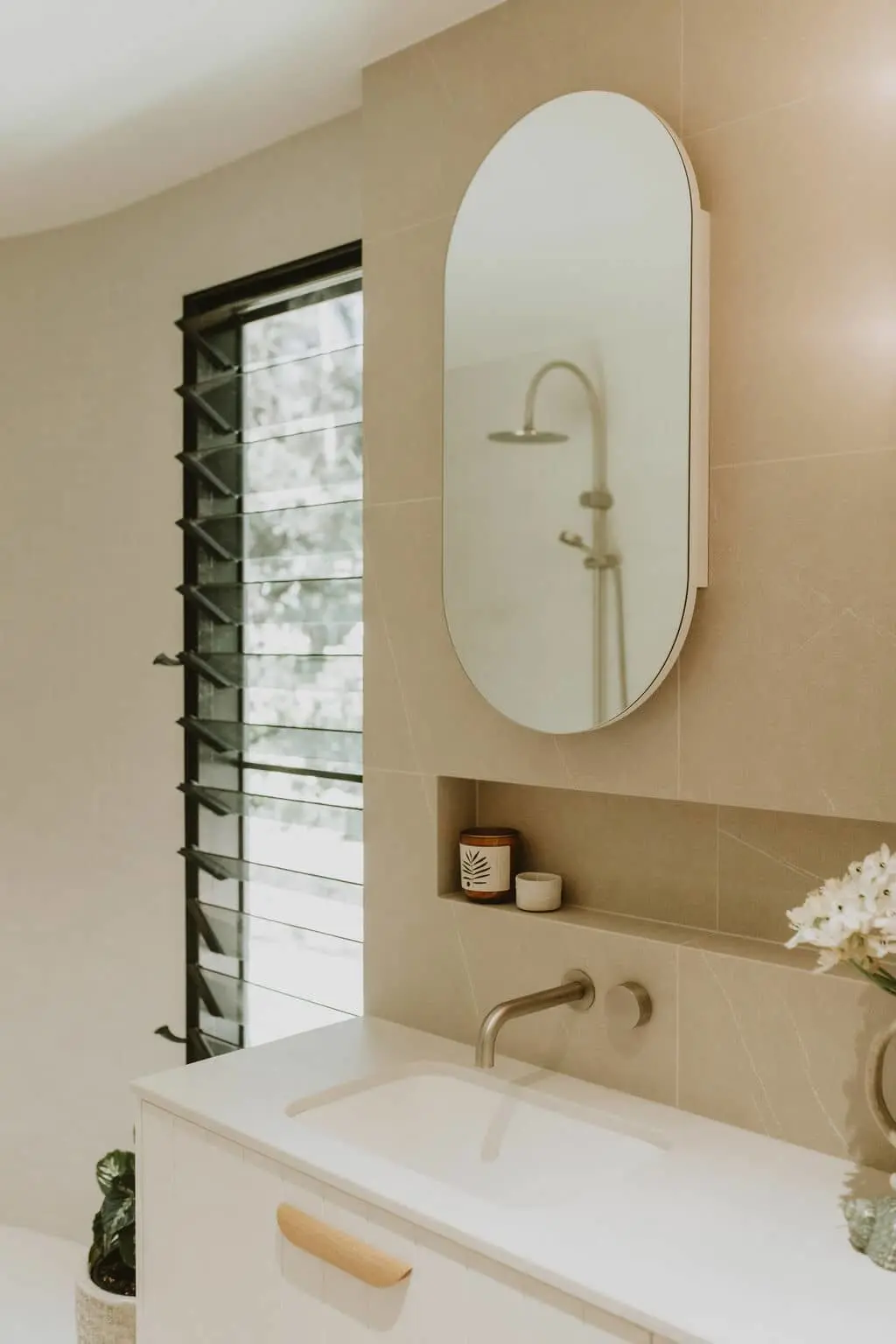 Modern bathroom with oval mirror, white sink, beige tiled walls, louvered window, and small wall niche holding candles.