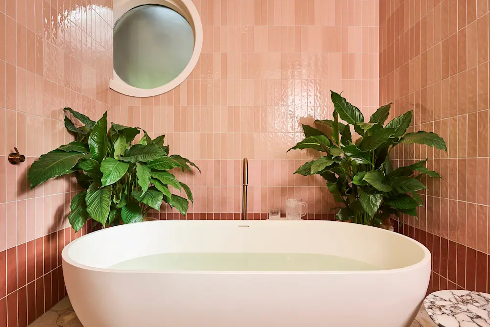 Modern bathroom with pink tiled walls, white freestanding bathtub, lush green plants, and a round window.