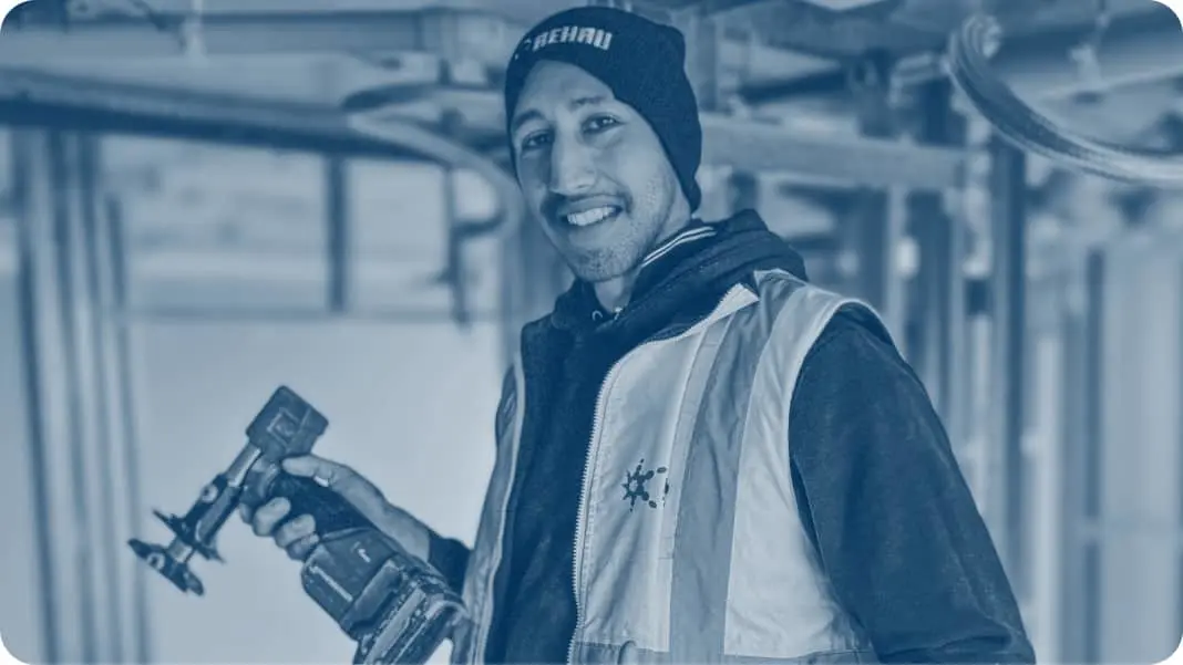 Person in beanie and work vest holding a power tool in an industrial setting, tinted in blue.