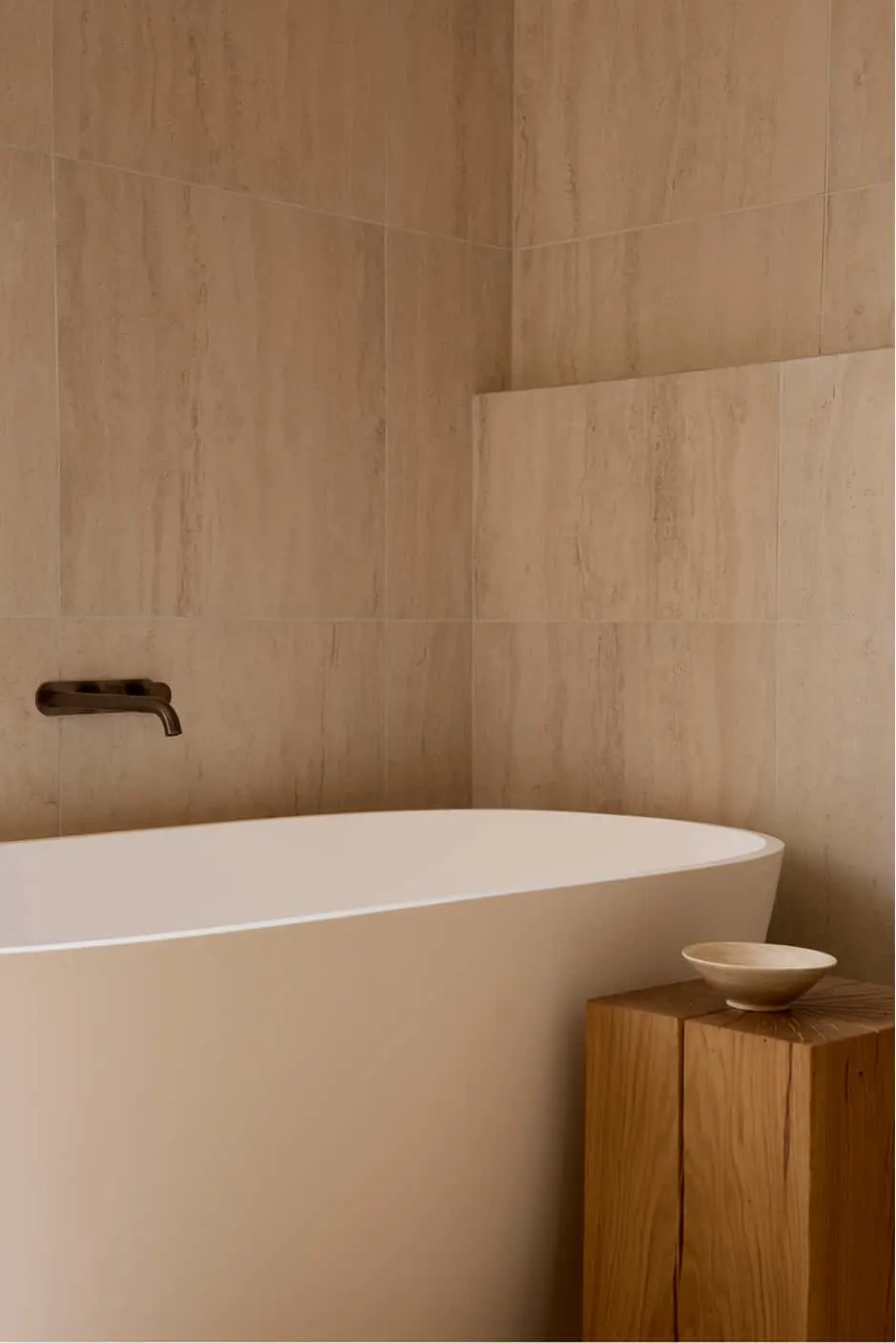 White freestanding bathtub against beige stone-tiled walls, with a dark bronze wall-mounted faucet and a wooden block side table.