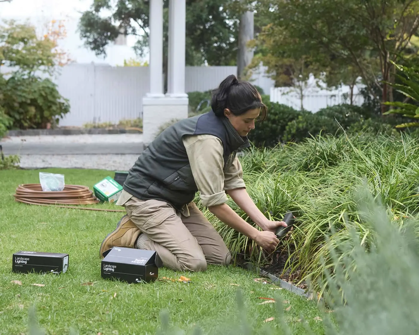 Person installing landscape lighting in garden, kneeling on grass with equipment boxes nearby in a residential yard.