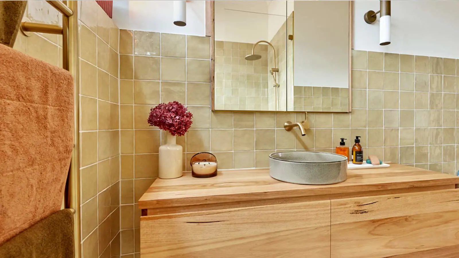 Modern bathroom with beige tiles, wooden vanity, round basin, gold fixtures, and red flowers in a white vase.