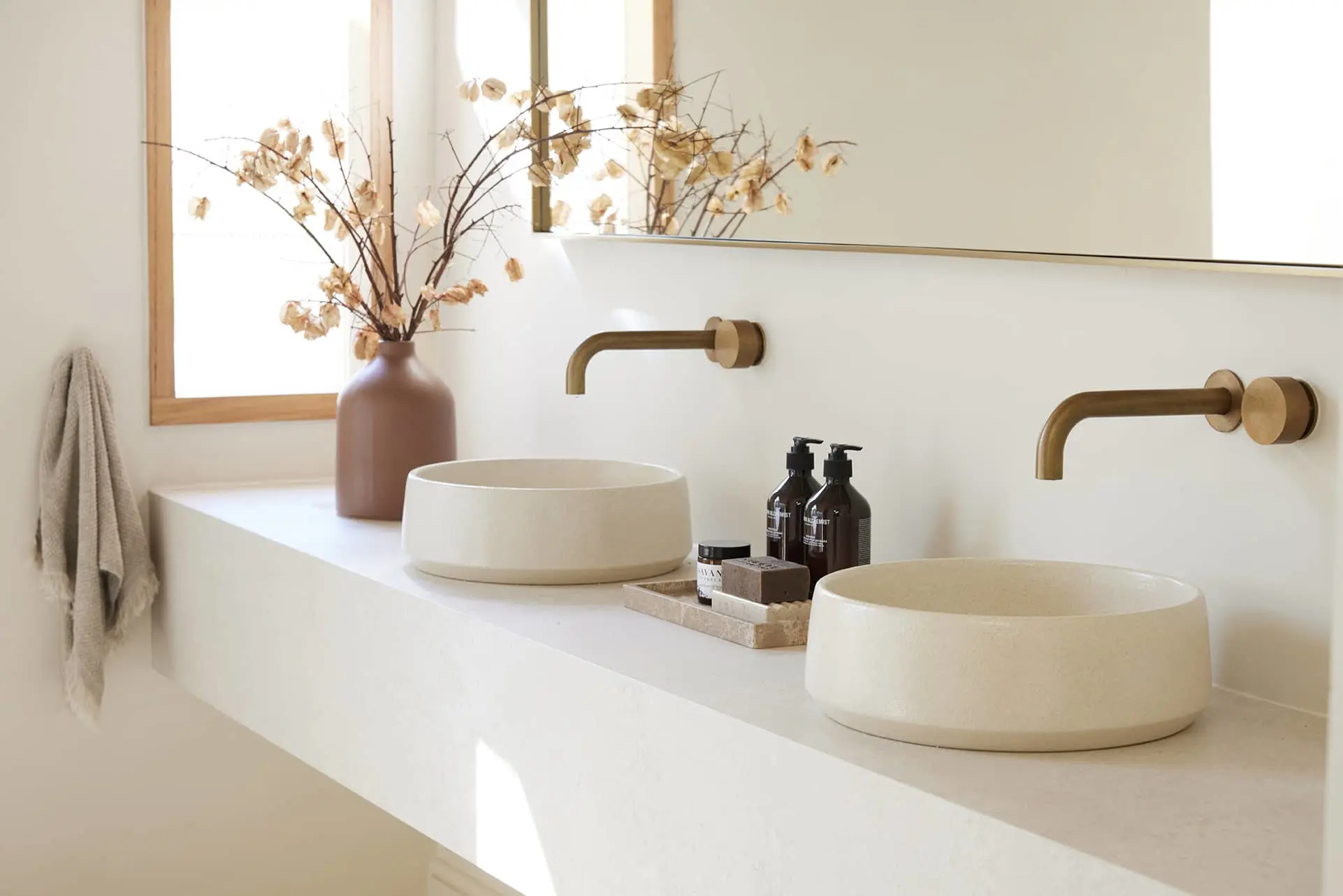 Modern bathroom with two white vessel sinks, brass wall-mounted faucets, and a terracotta vase with dried flowers.
