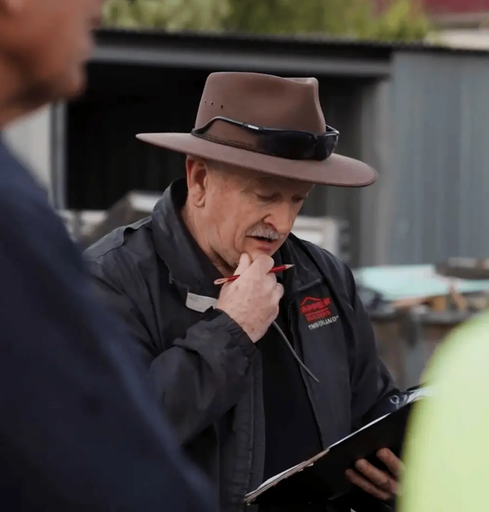 Person in brown hat and black jacket holding a pen to their chin while looking at documents in an outdoor setting.