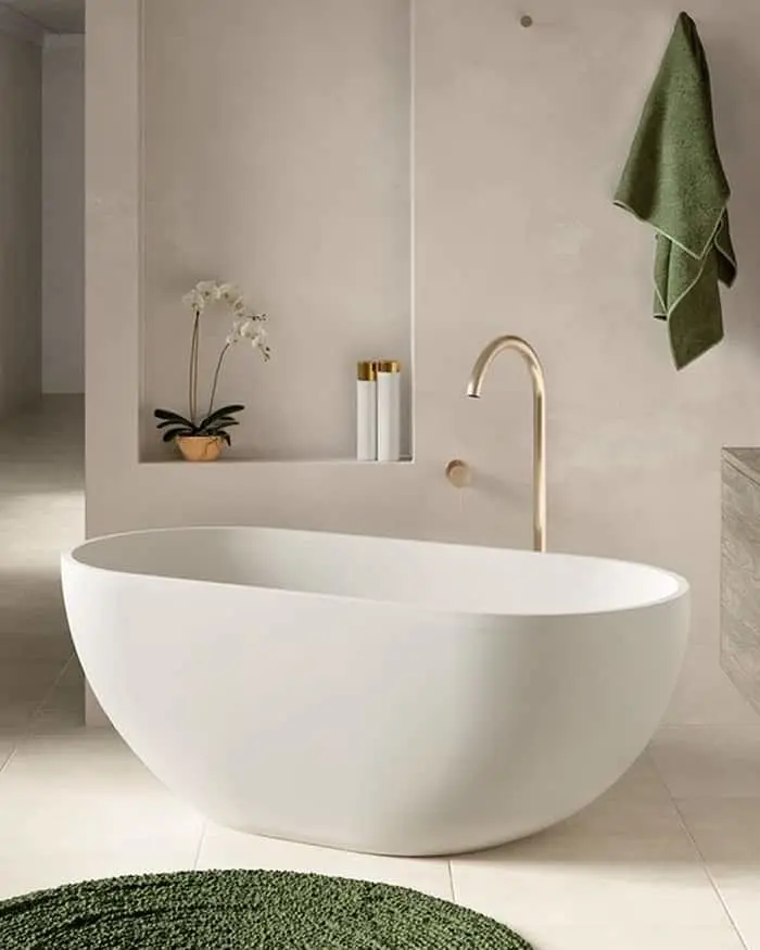 Modern white freestanding bathtub with brass faucet in minimalist bathroom featuring green towel and orchid plant.