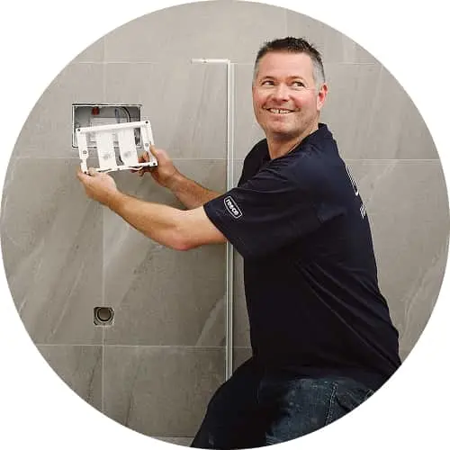Person in navy shirt installing a bathroom fixture on gray tiled wall, smiling while working