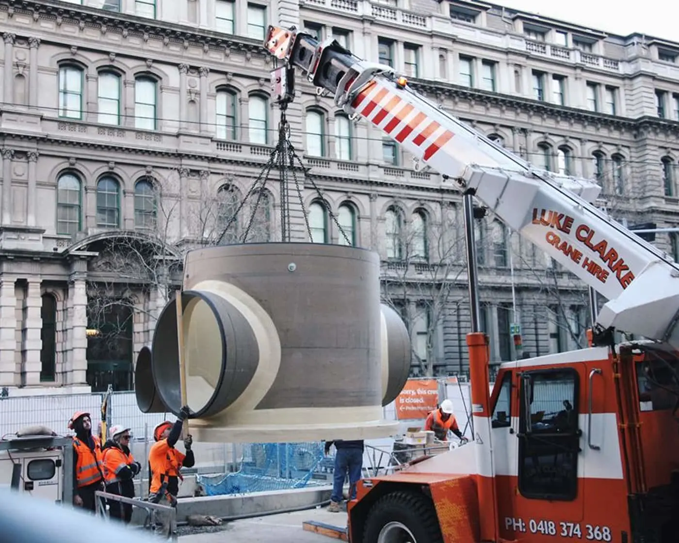 Construction workers in orange vests using a crane to install a large concrete pipe section in front of a historic building.