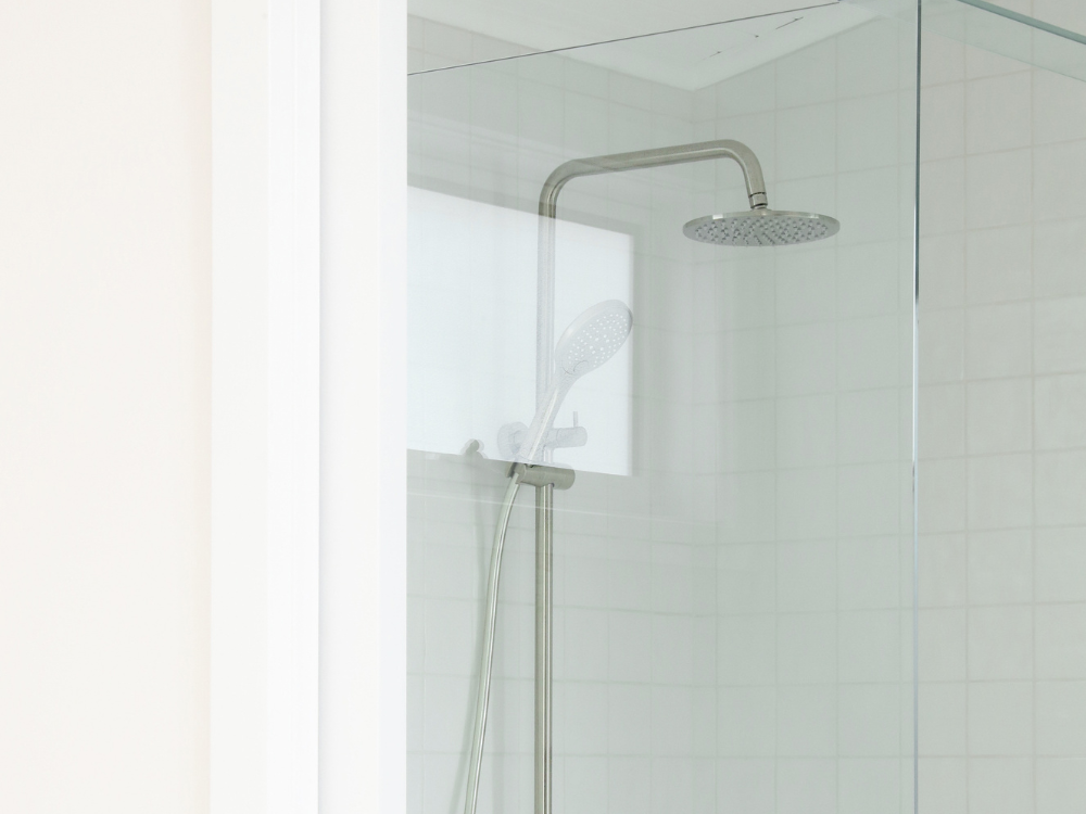 Modern shower stall with white tile walls, fixed rainfall showerhead, and handheld shower attachment in brushed metal finish.