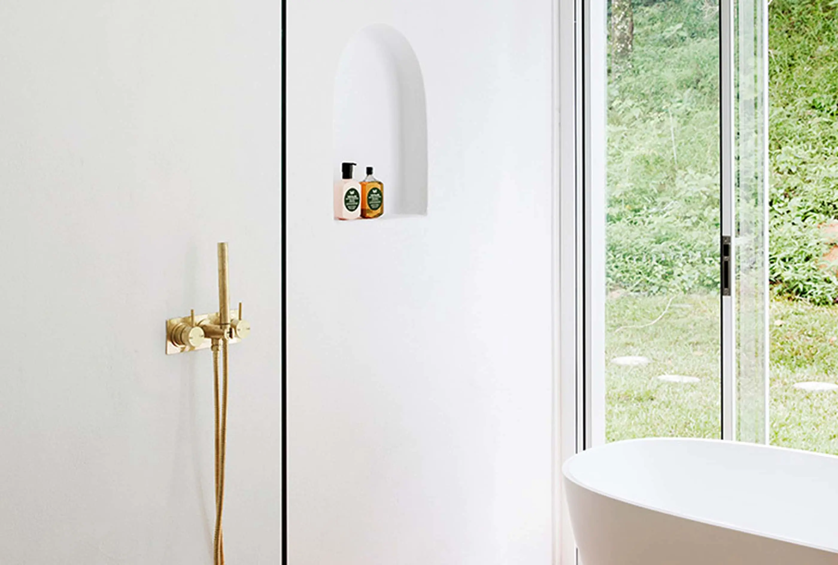 Modern white bathroom with brass fixtures, wall niche for toiletries, and large windows overlooking green garden.