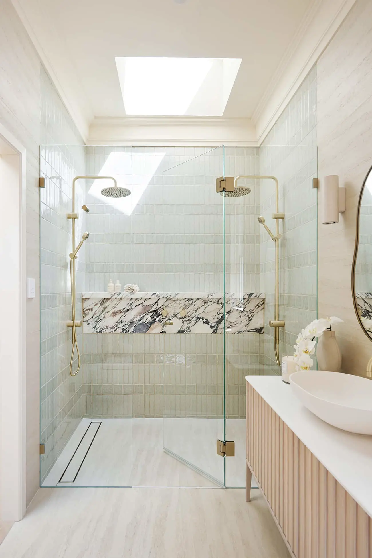 Modern bathroom with glass shower, pale green tiles, marble accent shelf, wooden vanity, and skylight providing natural light.