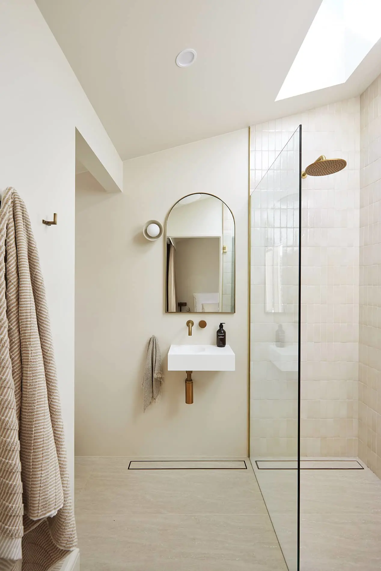 Modern minimalist bathroom with white walls, glass shower enclosure, small sink, arched mirror, and brass fixtures.