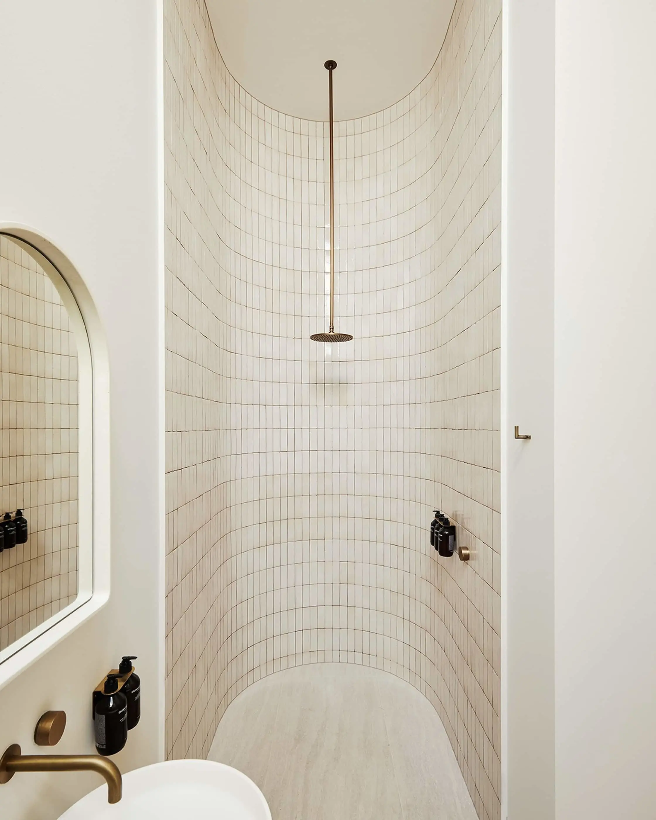 Minimalist curved shower with cream tile walls, brass rainfall showerhead, and partial view of sink with black toiletries.