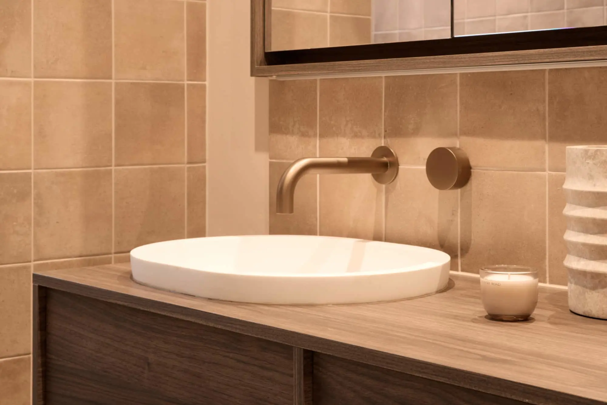 Modern bathroom with white vessel sink, brass wall-mounted faucet, wooden vanity, and beige tiled walls.