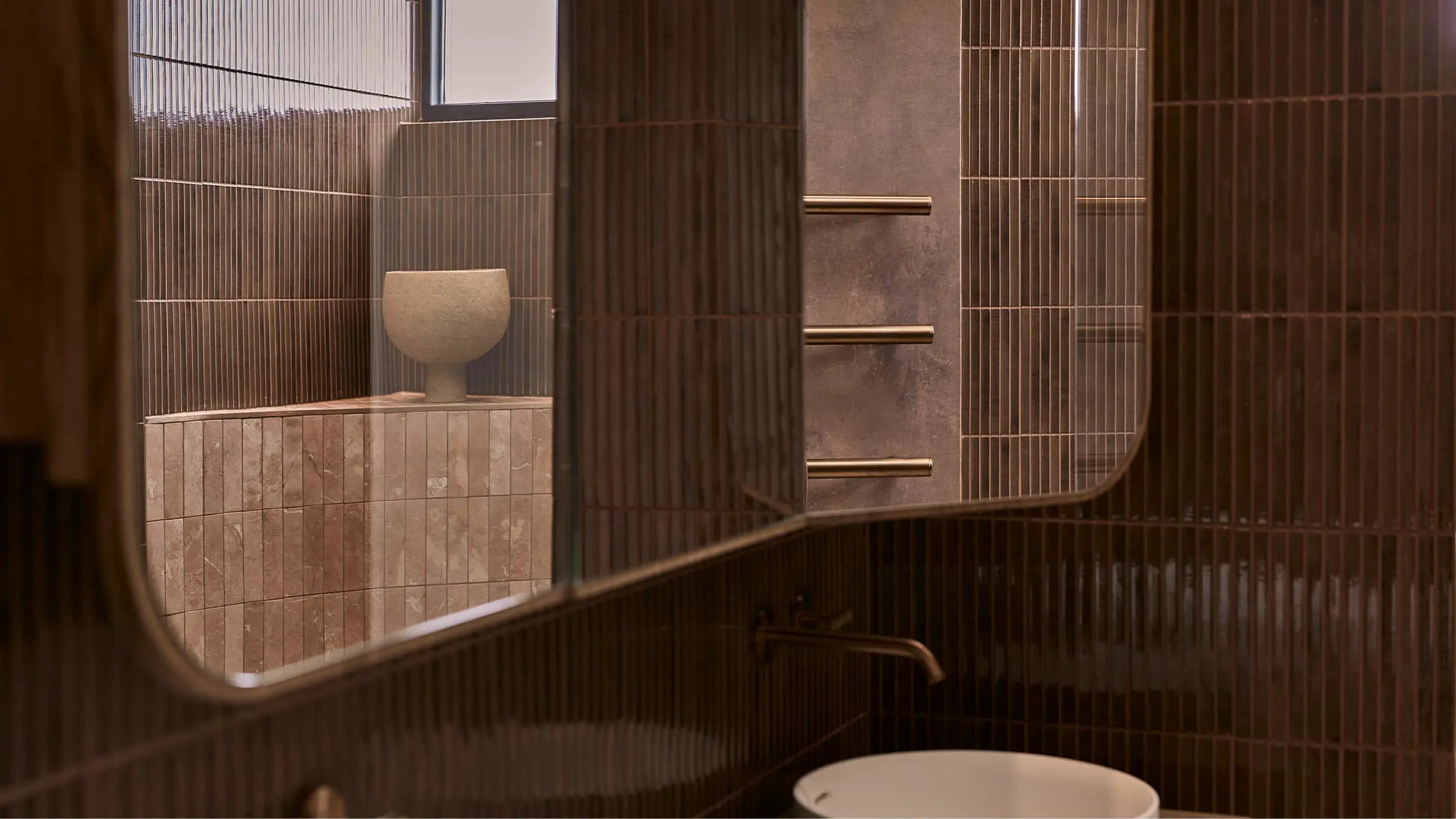 Modern bathroom with dark vertical tiles, curved mirror reflecting a beige vase on stone ledge, and brass fixtures.