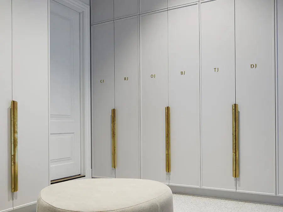 Modern white wardrobe with gold handles and initials, featuring a round cream ottoman in a minimalist dressing room.
