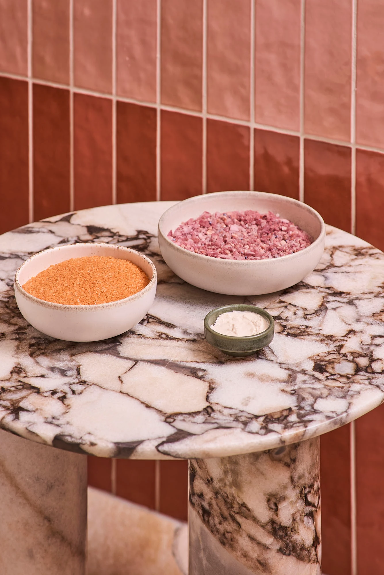 Two ceramic bowls with orange and pink bath salts and a small dish of cream on a marble table against terracotta tiles.