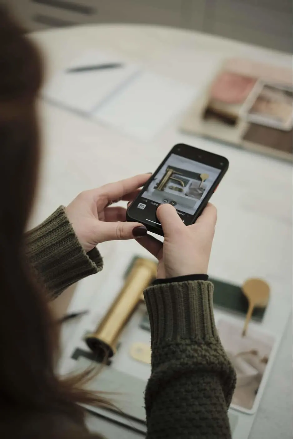 Person in green sweater looking at interior design image on smartphone while sitting at white table with materials.
