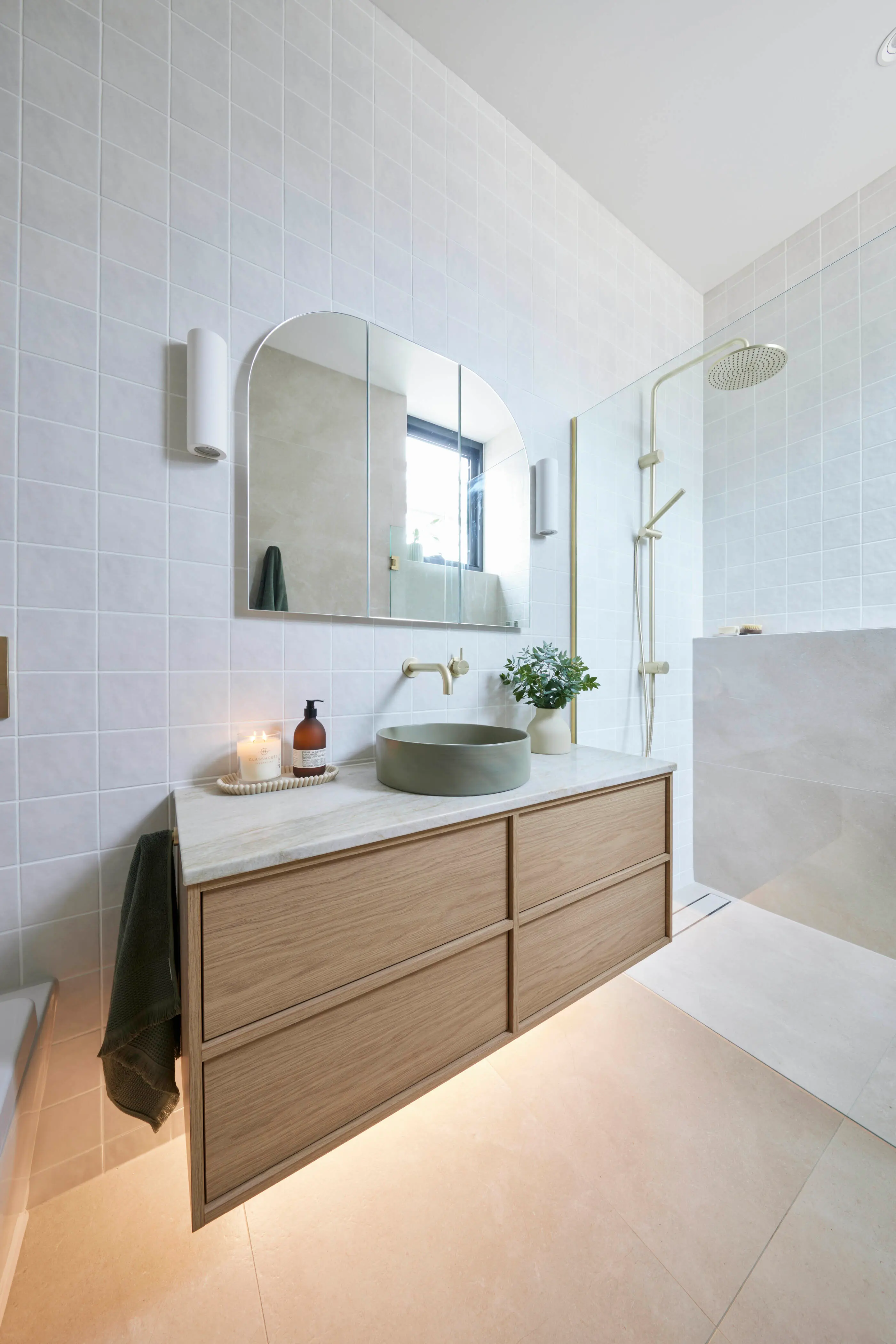 Modern bathroom with light blue tiles, wooden vanity, round green sink, curved mirror, and walk-in shower.
