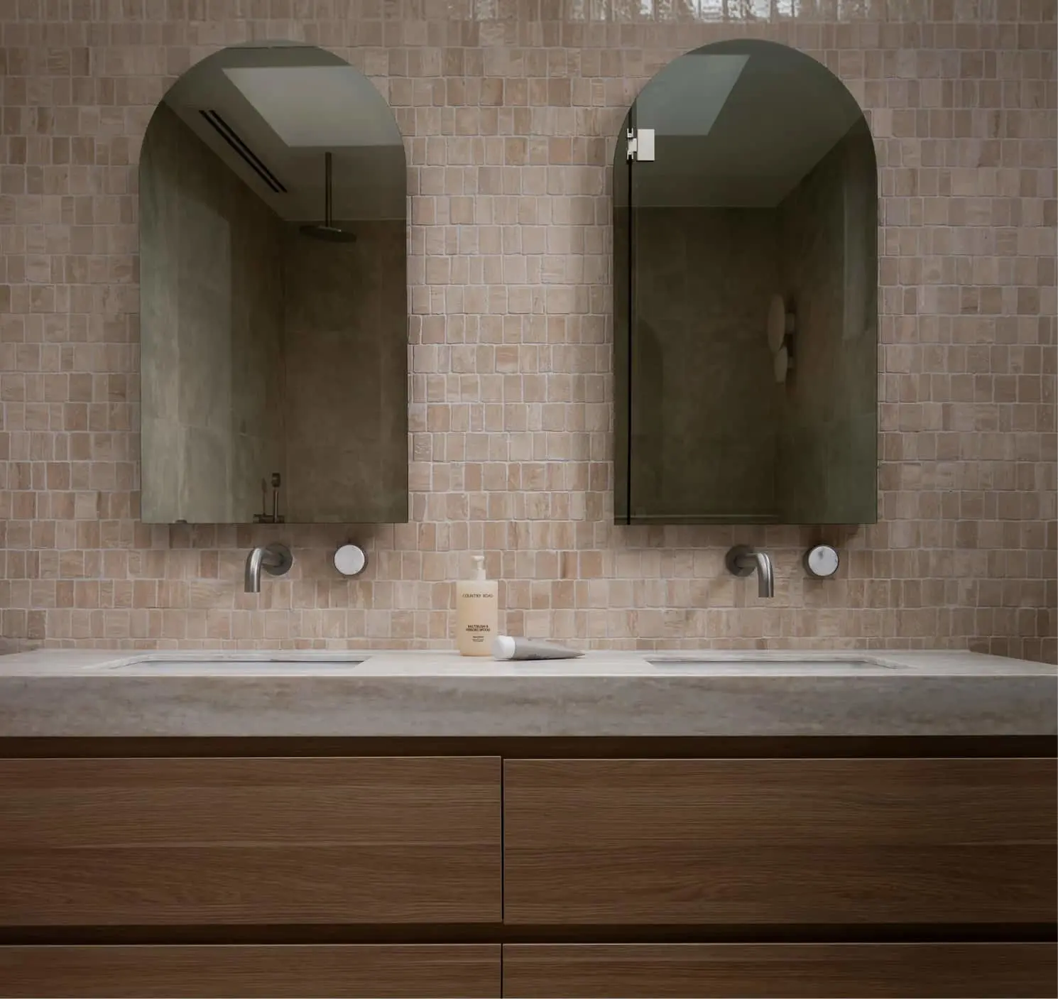 Double vanity bathroom with arched mirrors, beige tile wall, wall-mounted faucets, and wood vanity below.