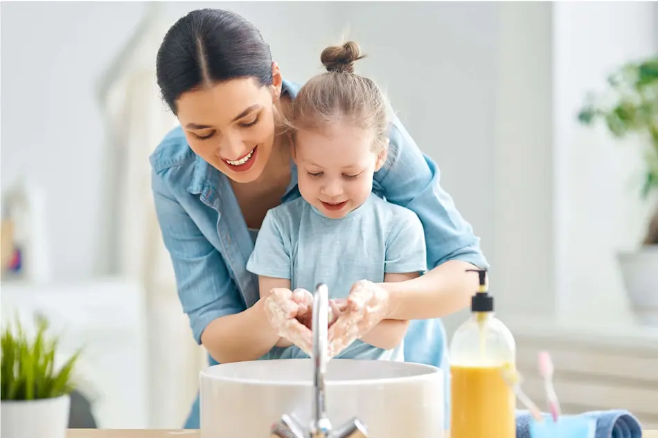 Adult and child in matching blue outfits washing hands together at a bathroom sink with soap dispenser nearby.