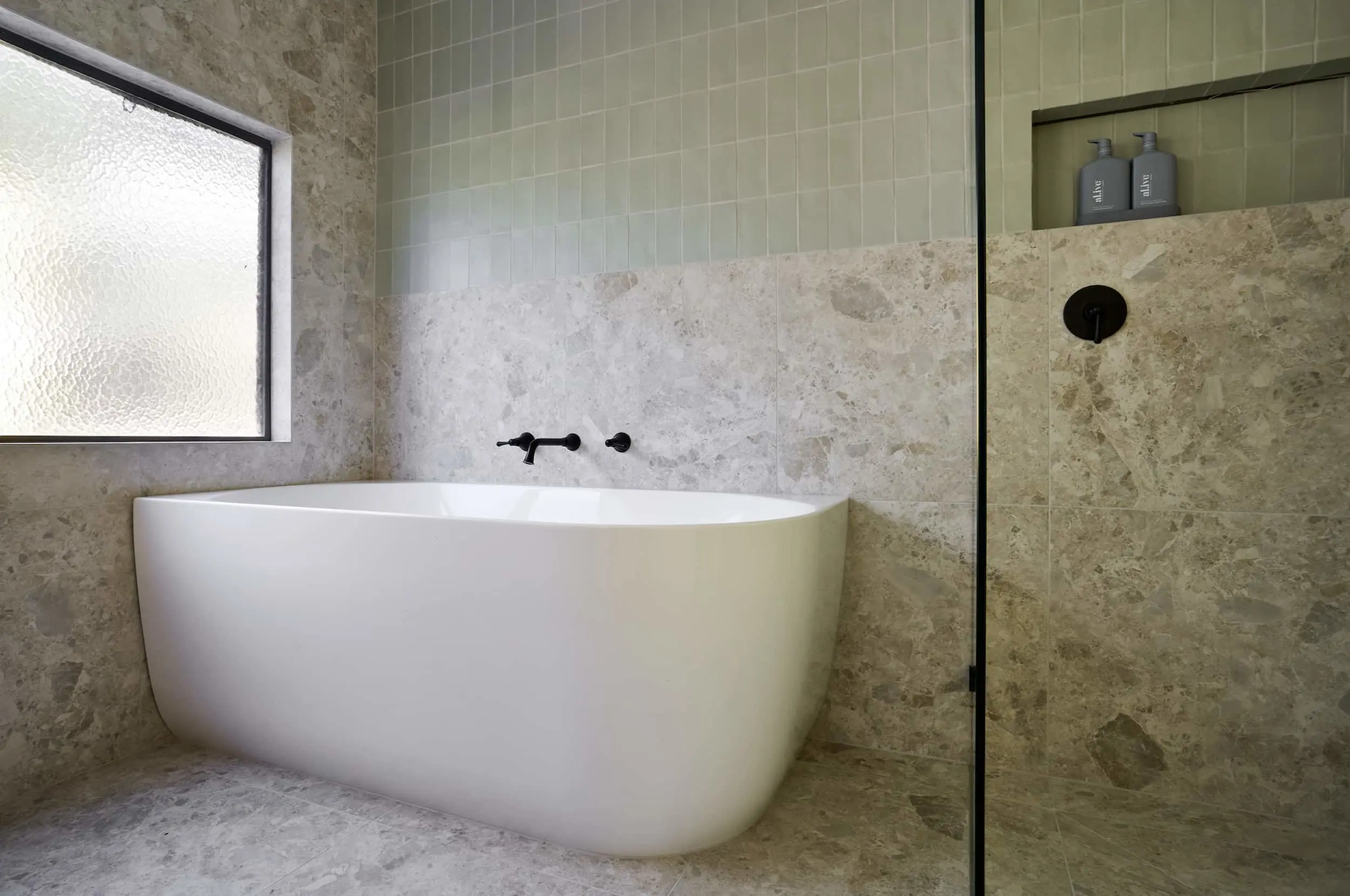 Modern bathroom with white freestanding tub, stone tile walls, frosted window, and glass shower with product niche.