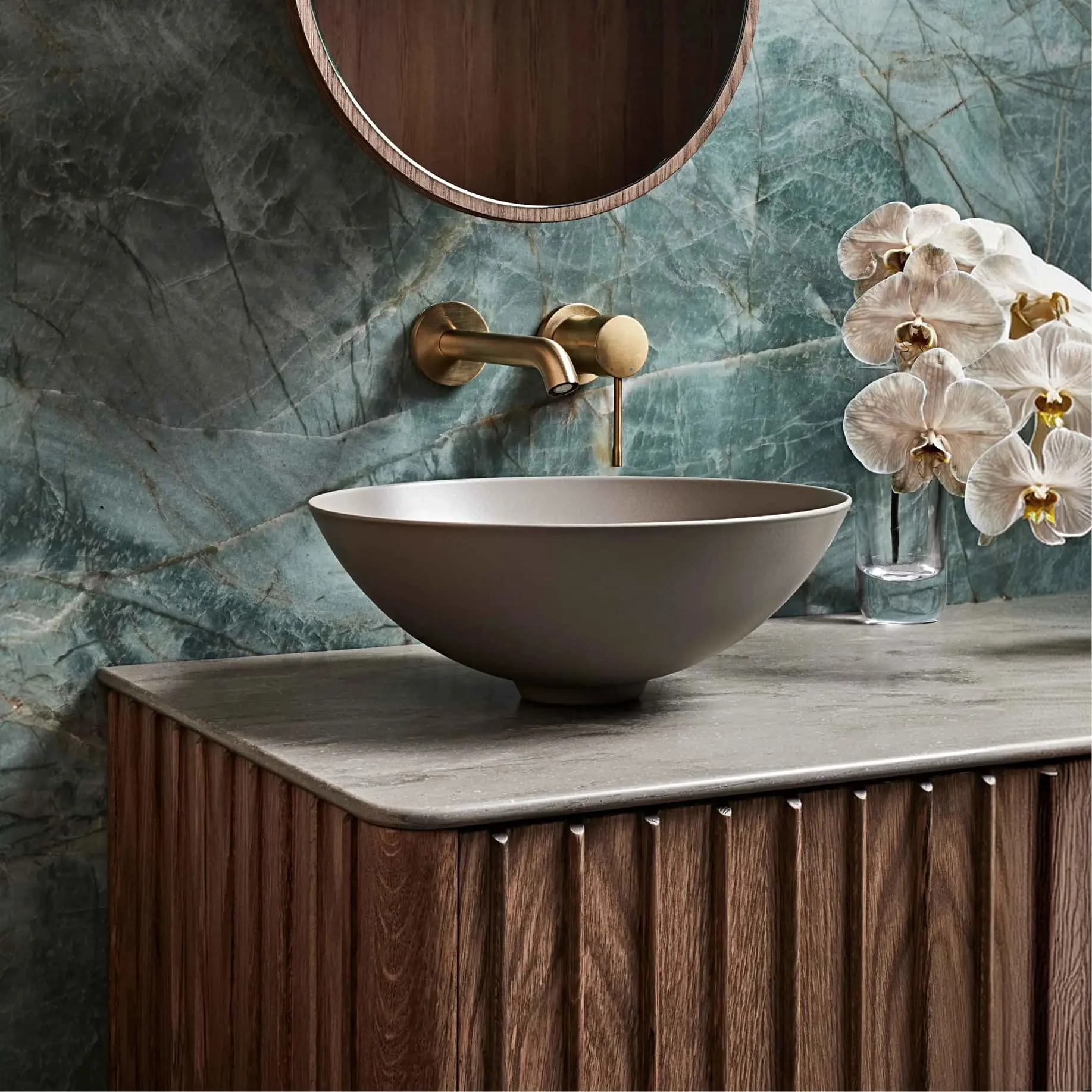 Modern bathroom with gray bowl sink on wood vanity, brass faucet, round mirror, and white orchids against blue marble wall.