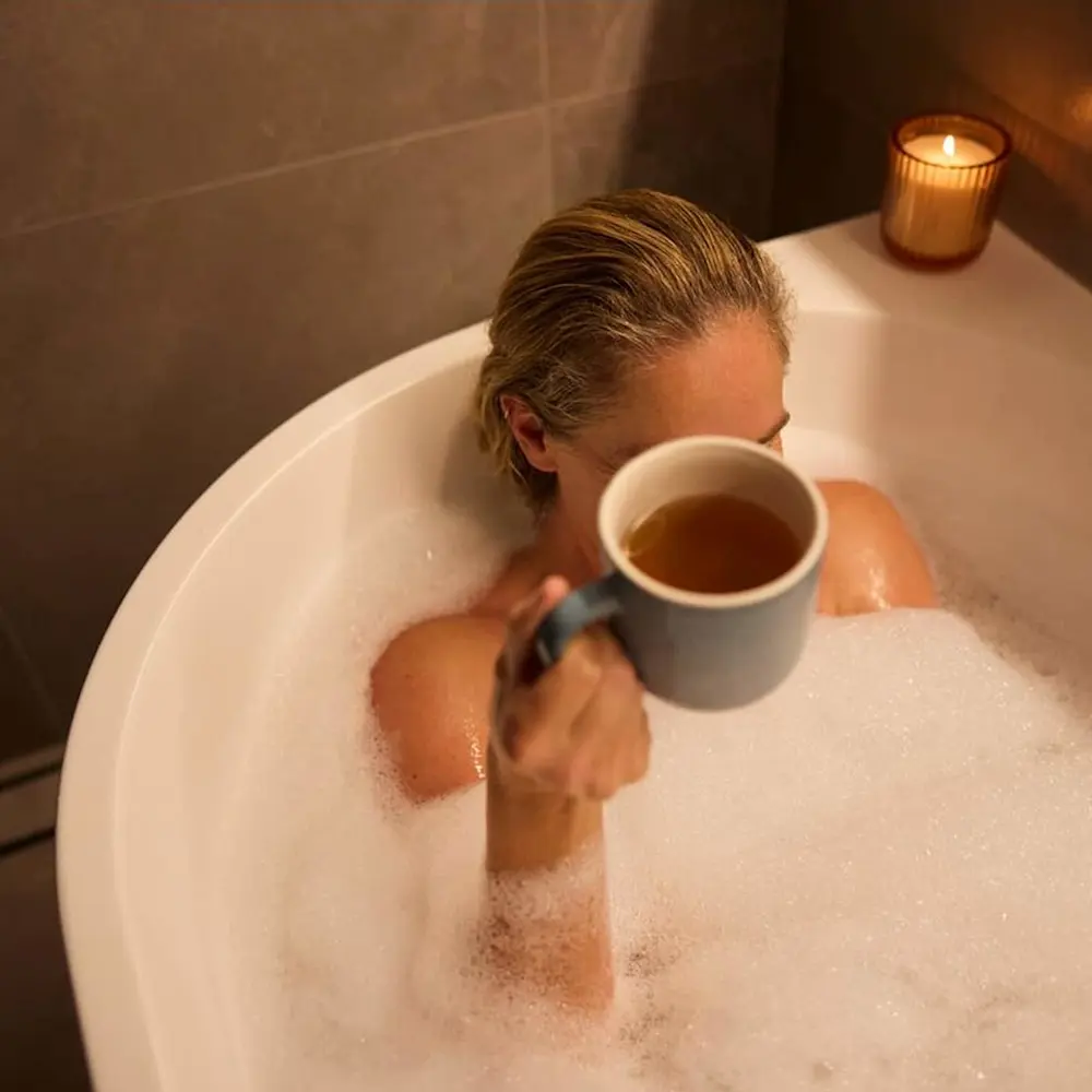 Person relaxing in bubble bath holding blue mug with tea, lit candle on bathtub edge in dim bathroom.