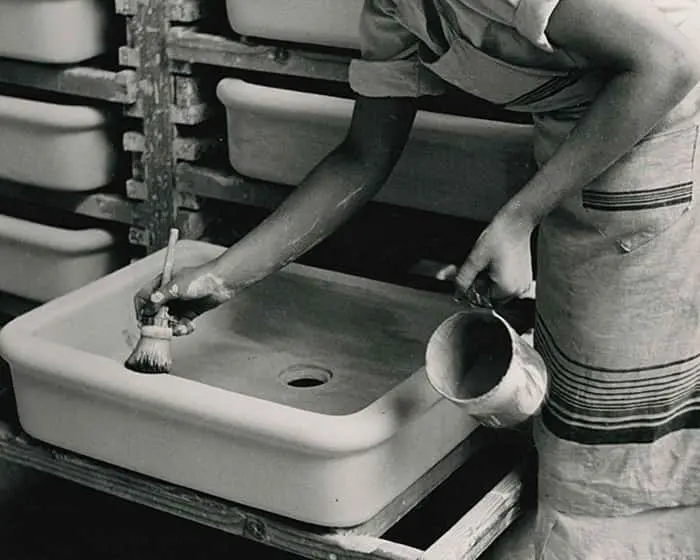 Person in apron applying glaze to ceramic sink in pottery workshop, with shelves of unfired pieces in background.