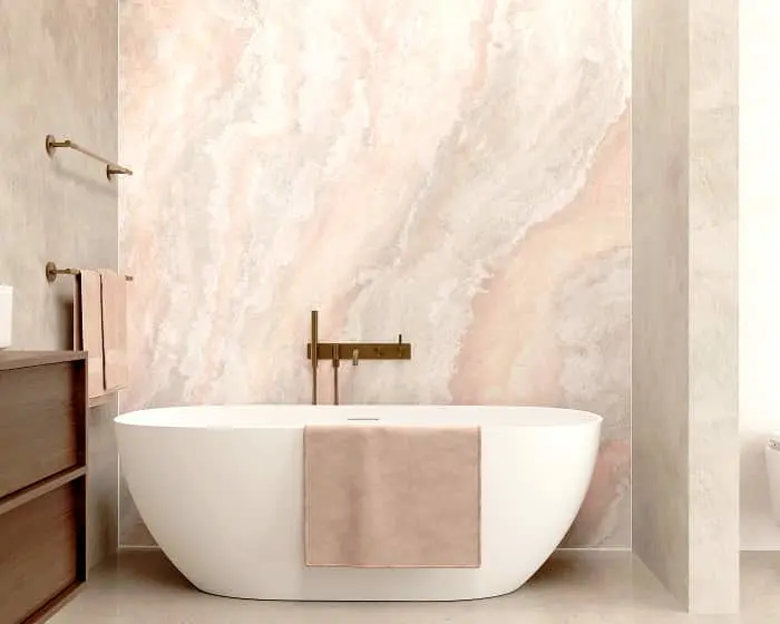 Modern bathroom with white freestanding tub against pink marble wall, brass fixtures, and pink towels.