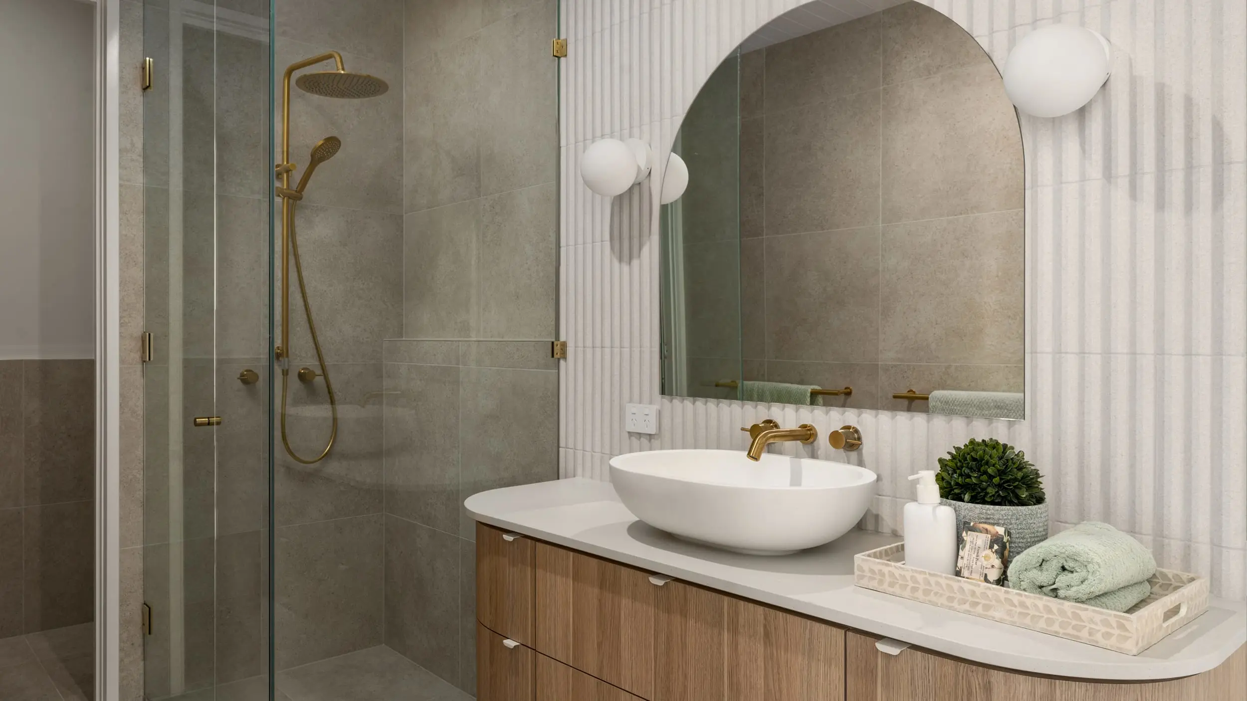 Modern bathroom with glass shower, white vessel sink, wooden vanity, arched mirror, and gold fixtures.