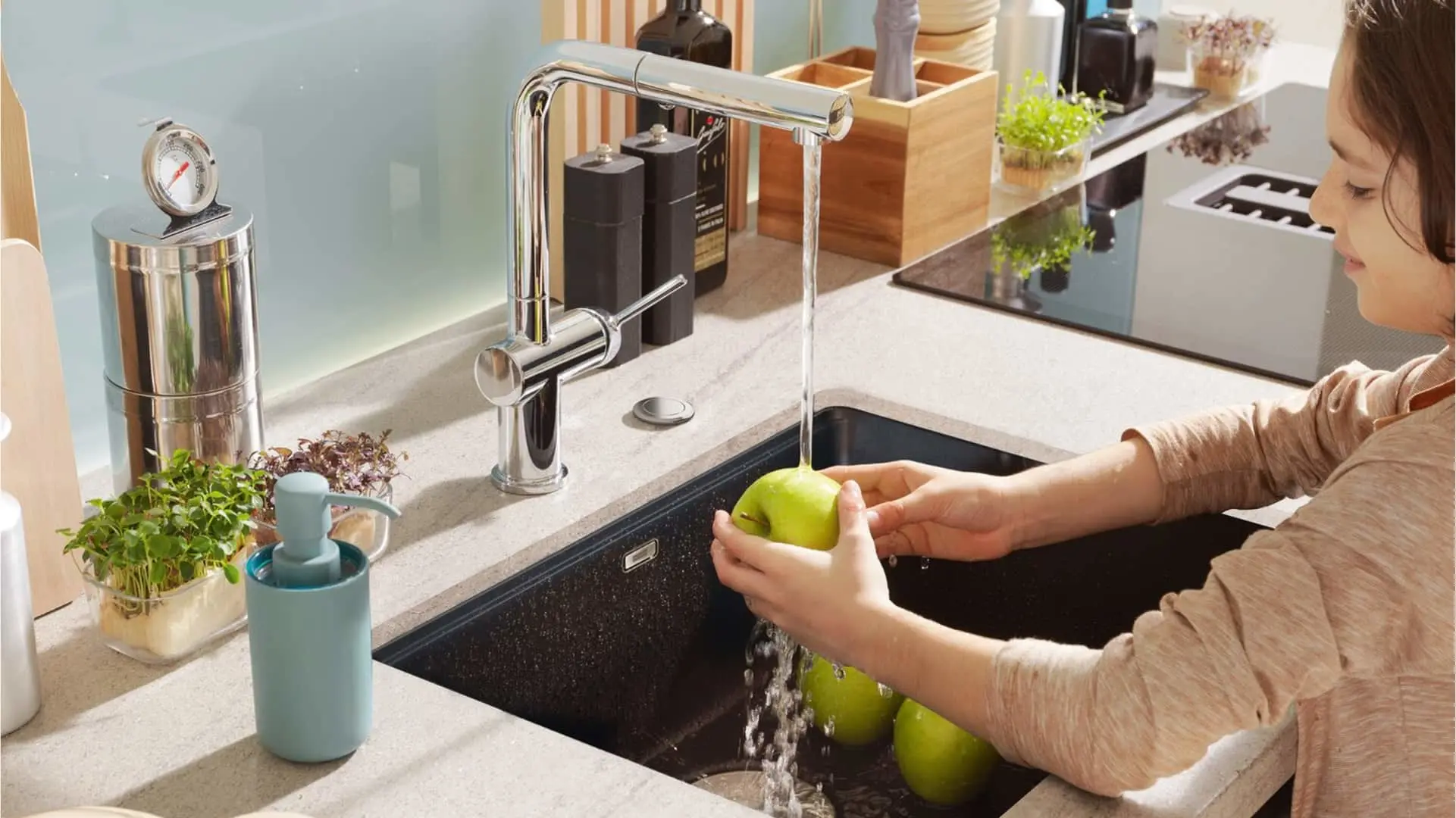 Person washing green apples in modern kitchen sink with chrome faucet, stone countertop, and small plants nearby.