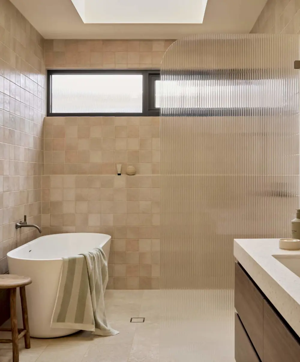 Modern bathroom with beige tiled walls featuring a freestanding white bathtub with towel and wall-mounted faucet.