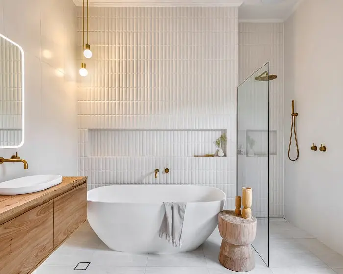 Modern white bathroom with freestanding tub, textured wall tiles, wooden vanity, and gold fixtures under pendant lights.