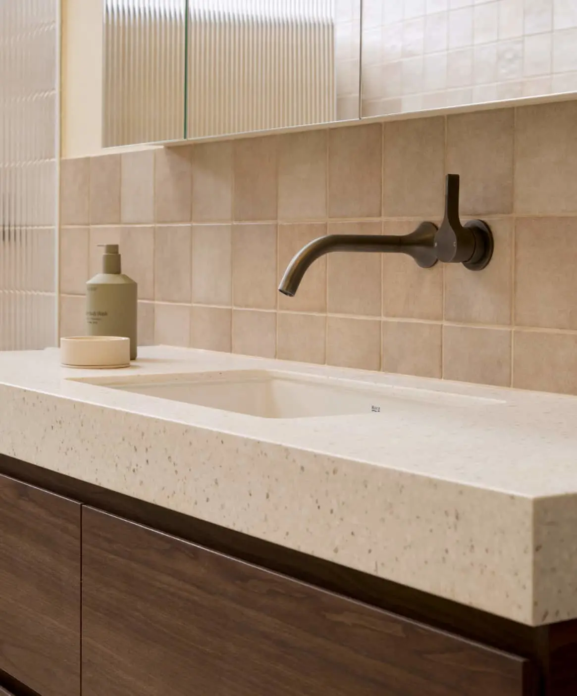 Modern bathroom sink with cream stone countertop, gunmetal wall-mounted faucet, beige tiled backsplash and soap dispenser.
