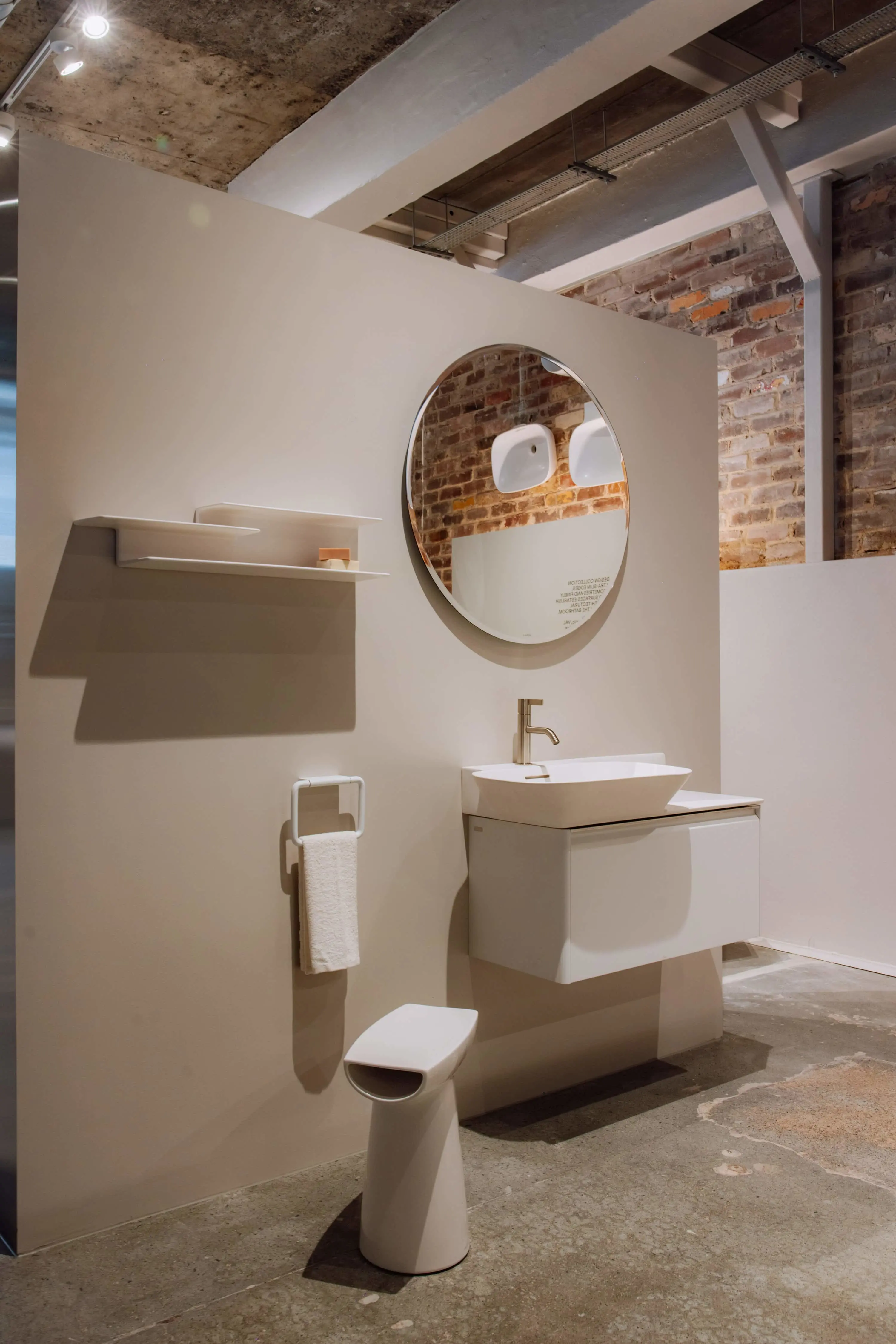 Modern bathroom display with white fixtures, round mirror, and exposed brick wall in an industrial-style showroom setting.