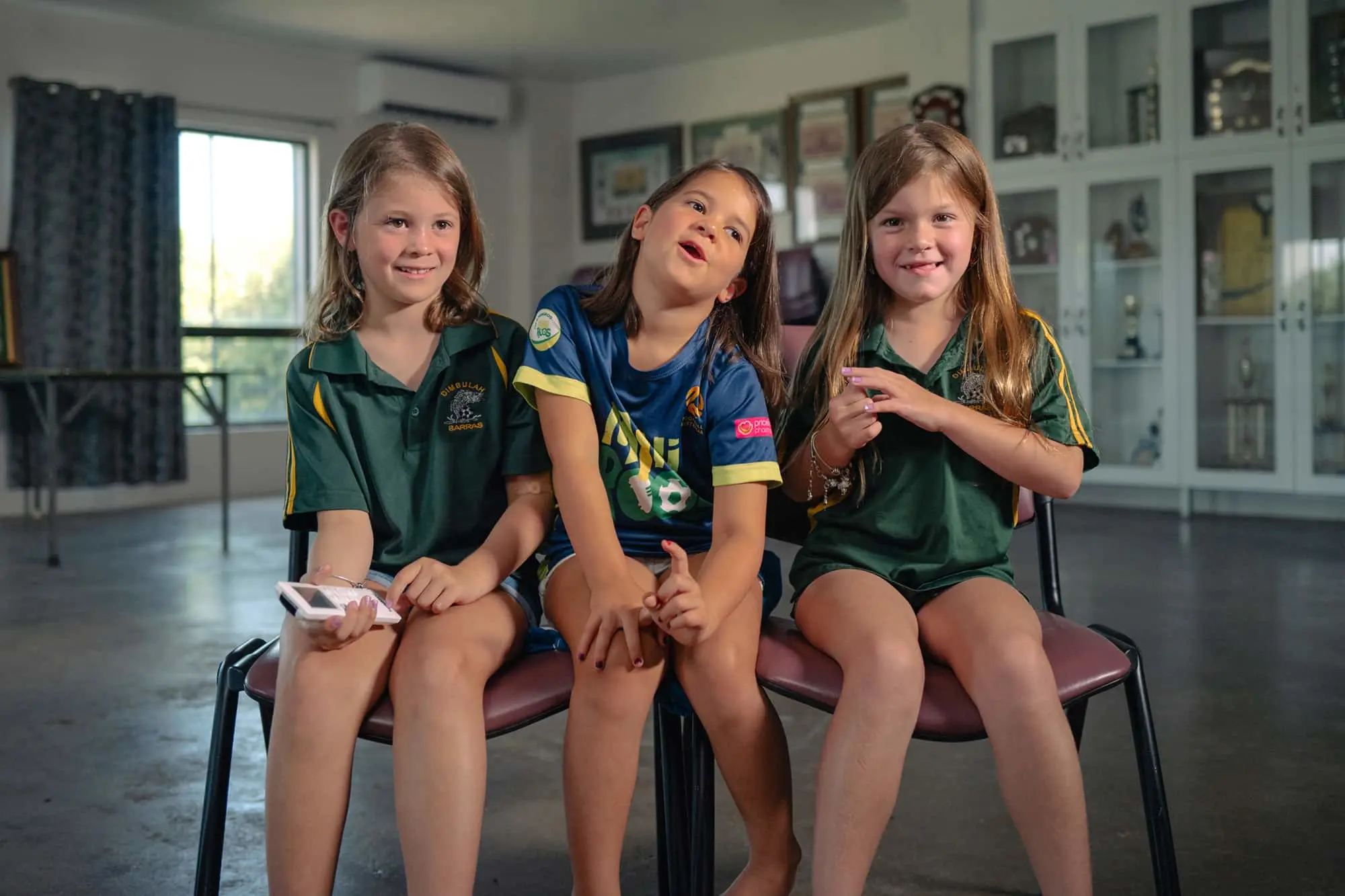 Three children sitting together on a bench, wearing green and blue sports uniforms in a bright indoor space.