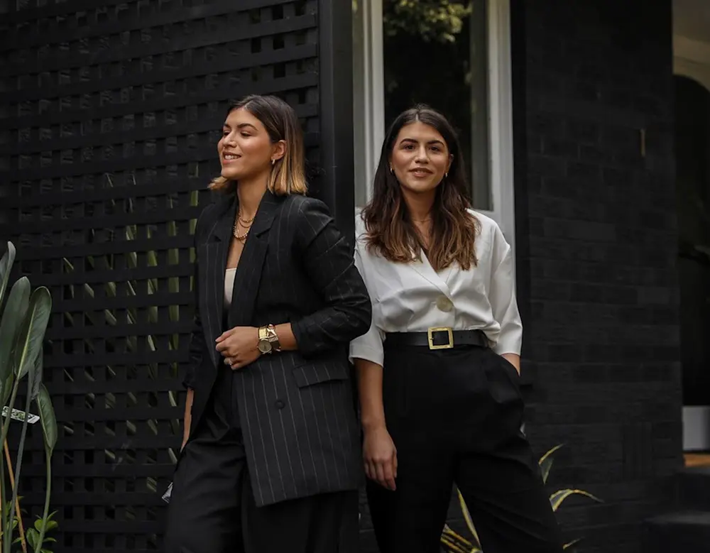 Two women in business attire standing against a black lattice wall, one in a pinstriped suit, one in white blouse and black pants.