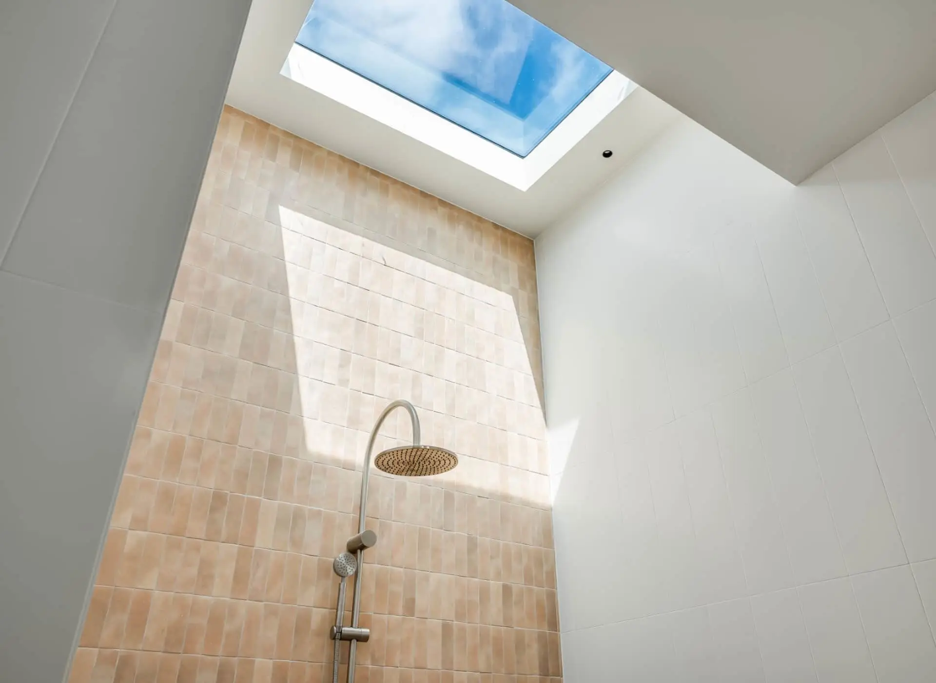 Modern shower with peach-colored tile wall, rainfall showerhead, and skylight showing blue sky above.