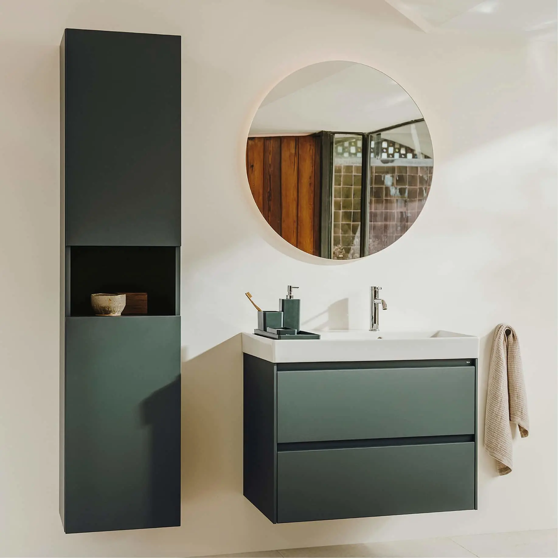 Modern bathroom with round mirror, white sink, green vanity cabinet, tall storage unit, and beige towel on white wall.