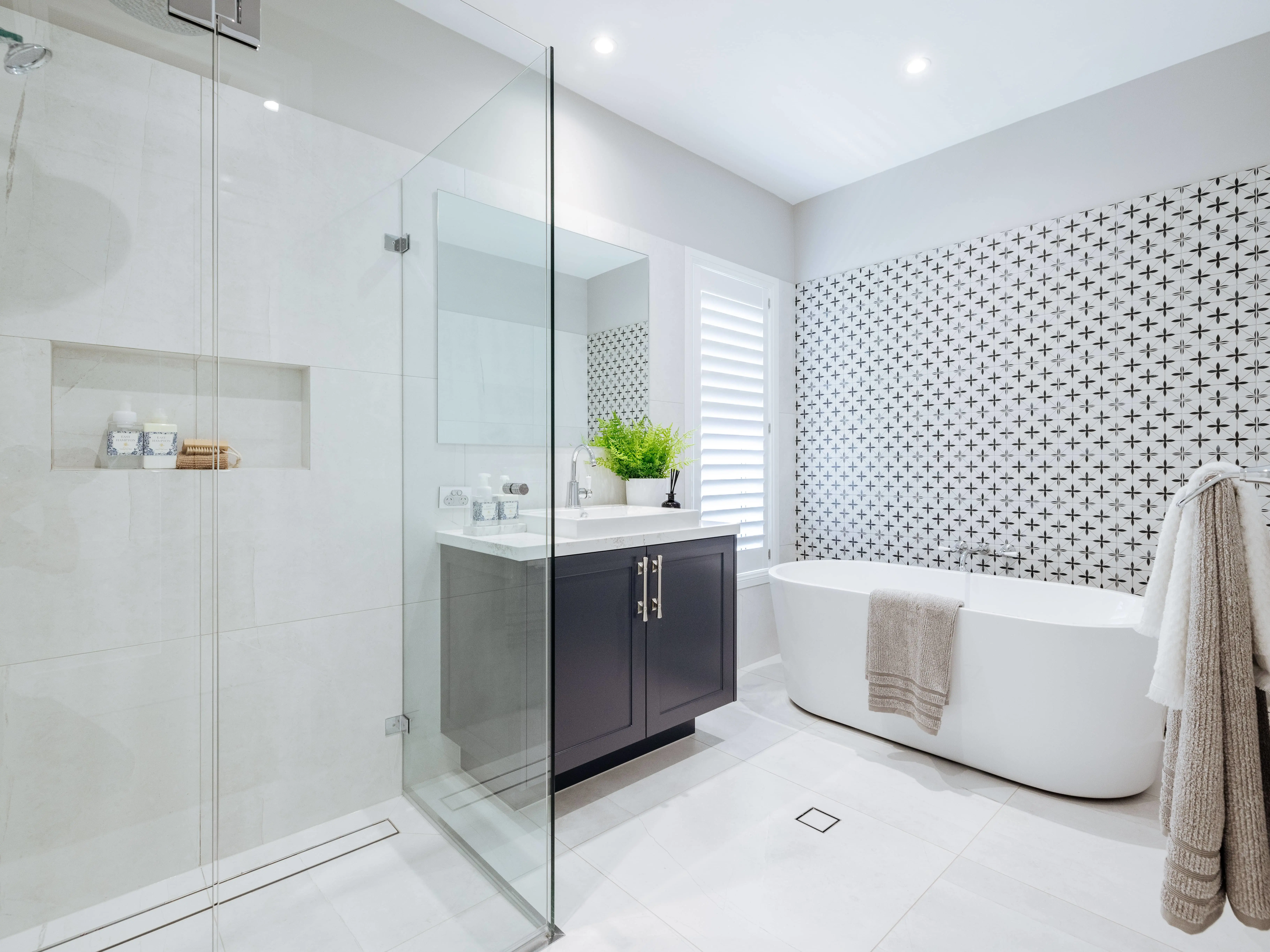 Modern bathroom with glass shower, dark vanity, white freestanding tub, and patterned accent wall.