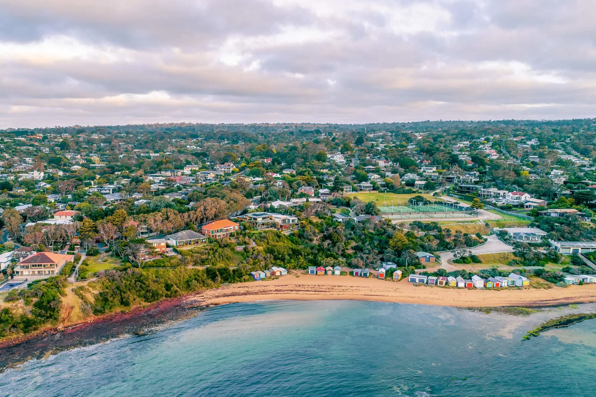 Aerial view of a coastal suburb with a sandy beach lined with colorful beach boxes, green trees, and residential houses.