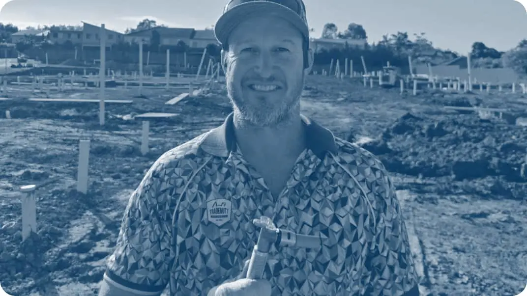 Person in patterned shirt and cap at construction site with foundation posts visible in background, blue-tinted image.