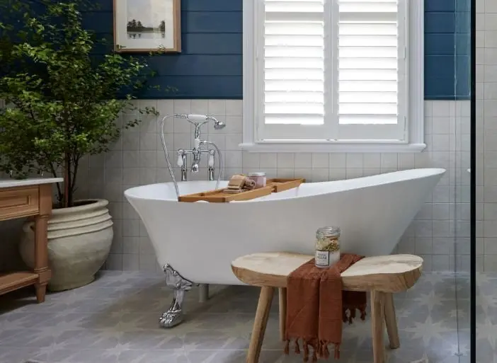 Elegant bathroom with white clawfoot tub, wooden stool, potted plant, and window with white shutters against blue wall.